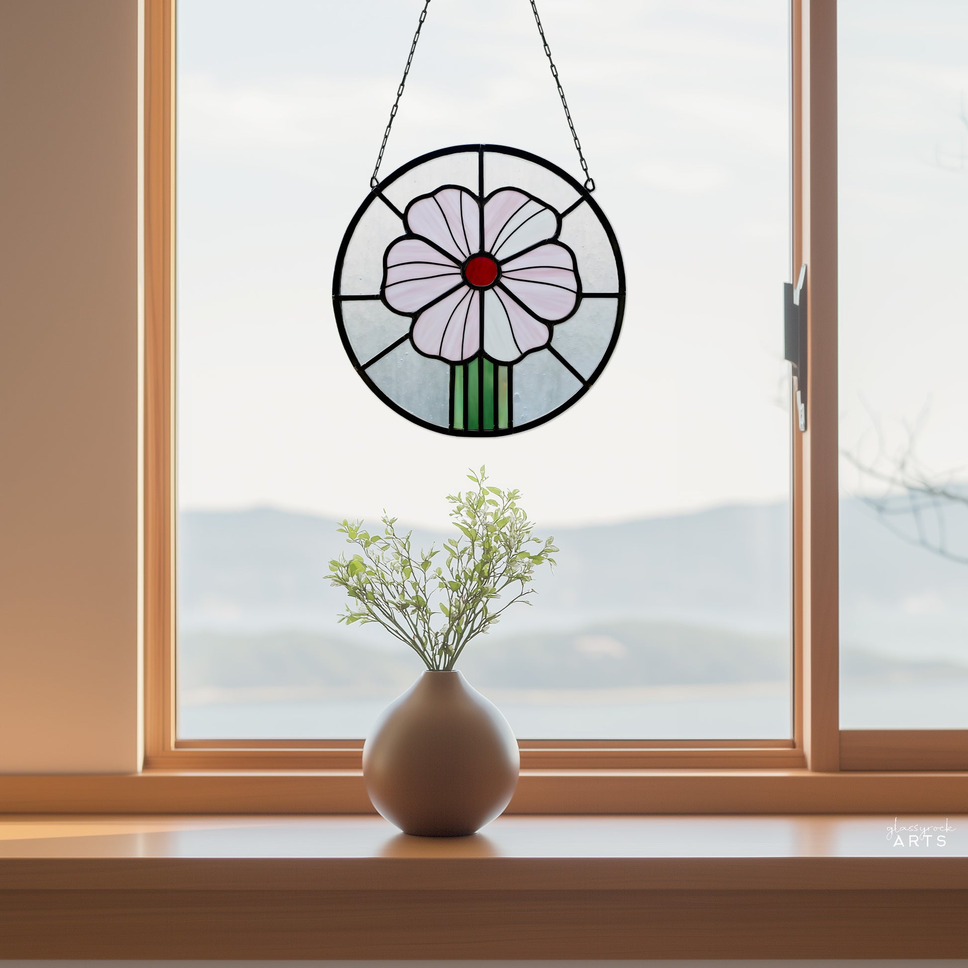 A round stained glass panel with a pink flower and red center, hanging in a window with a mountain background, with a vase on the windowsill. 