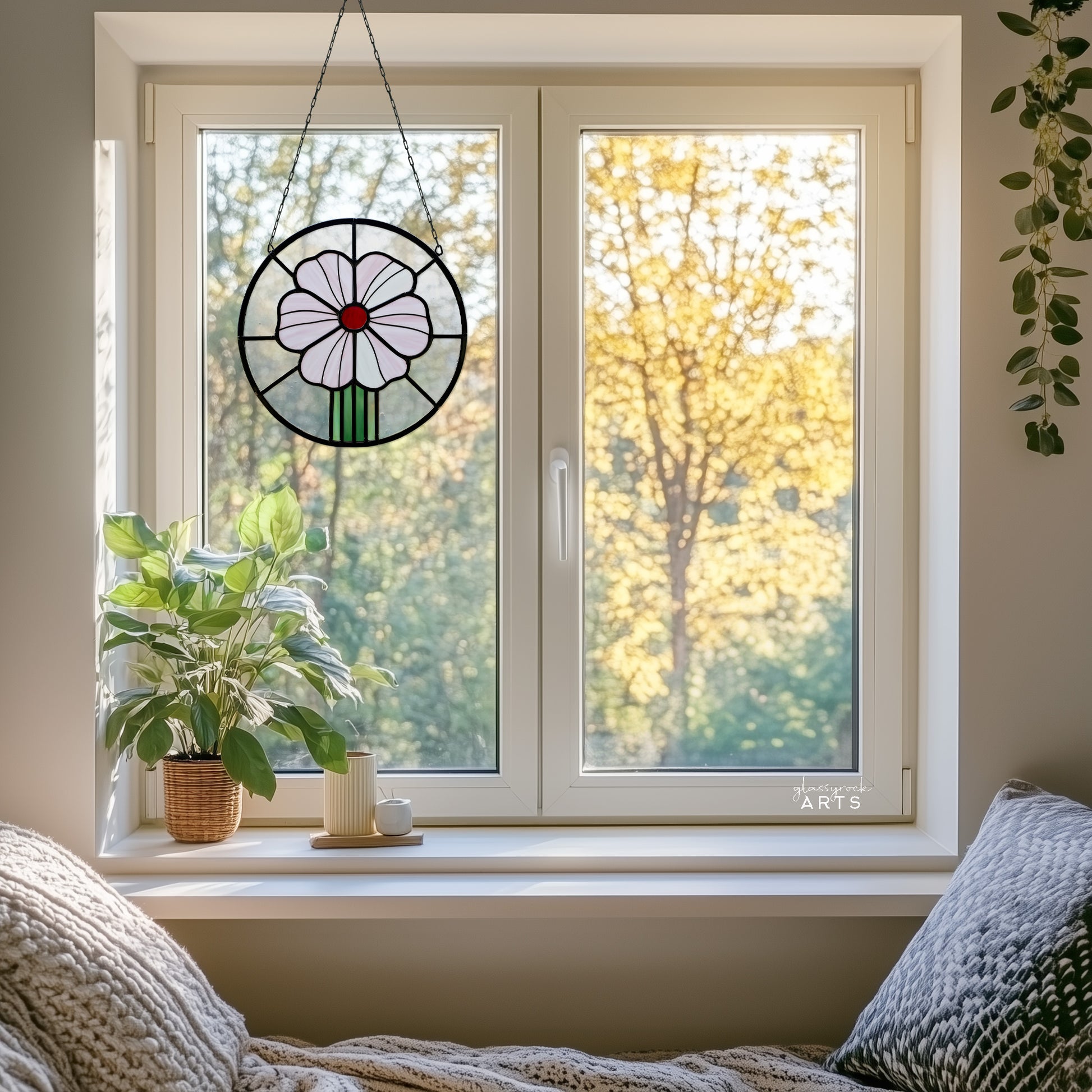 A round stained glass panel with a pink flower and red center, hanging in a window with a fall trees background, with a plant and vases on the windowsill. 