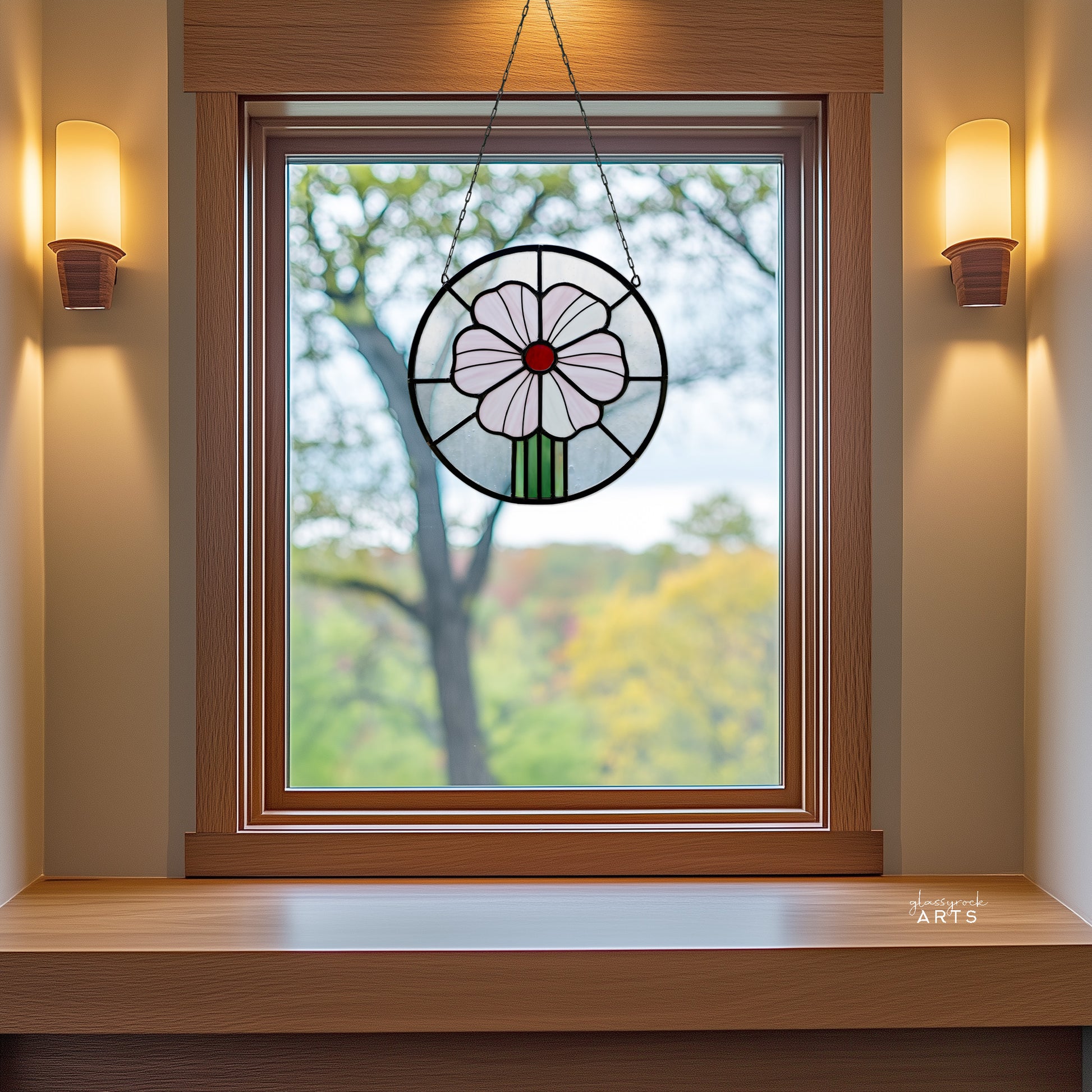 A round stained glass panel with a pink flower and red center, hanging in a window with a fall trees background.