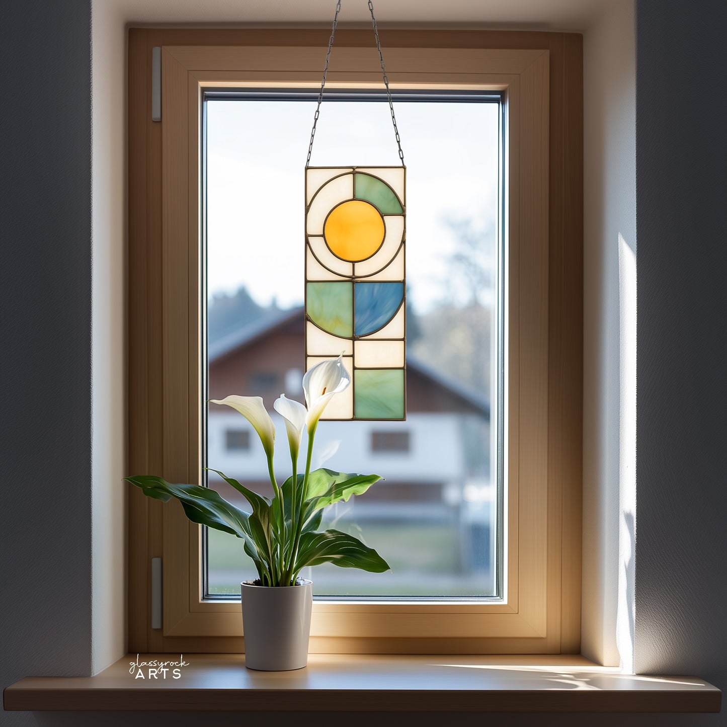 A yellow, white, blue, and green stained glass panel featuring a sun above and abstract straight and curvilinear shapes below. The panel is rectangular and tall. It hangs in a window with a house and trees behind it.