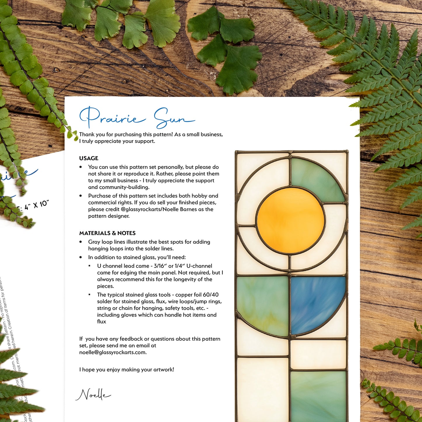 Two pages of the prairie sun stained glass pattern lying on a wood table with ferns.