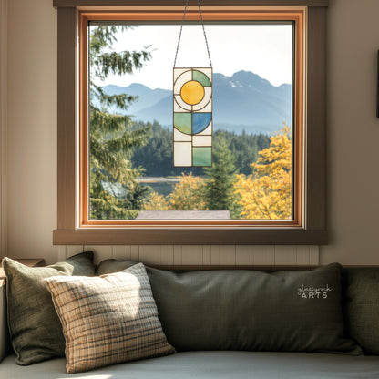 A yellow, white, blue, and green stained glass panel featuring a sun above and abstract straight and curvilinear shapes below. The panel is rectangular and tall. It hangs in a window with a lake and mountains behind it.