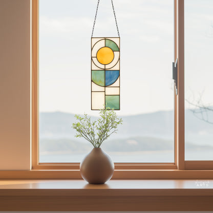 A yellow, white, blue, and green stained glass panel featuring a sun above and abstract straight and curvilinear shapes below. The panel is rectangular and tall. It hangs in a window with a lake and mountains behind it.