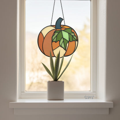 A pumpkin suncatcher, hanging in a window with fall foliage behind, and a small aloe plant on the windowsill.