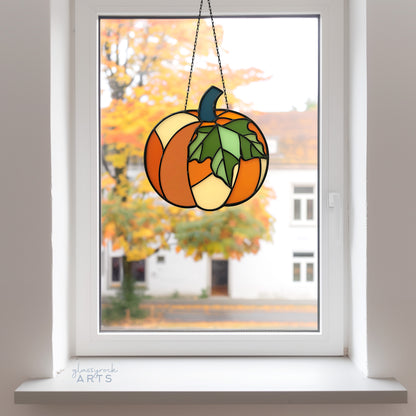 A pumpkin suncatcher, hanging in a window with fall foliage behind.