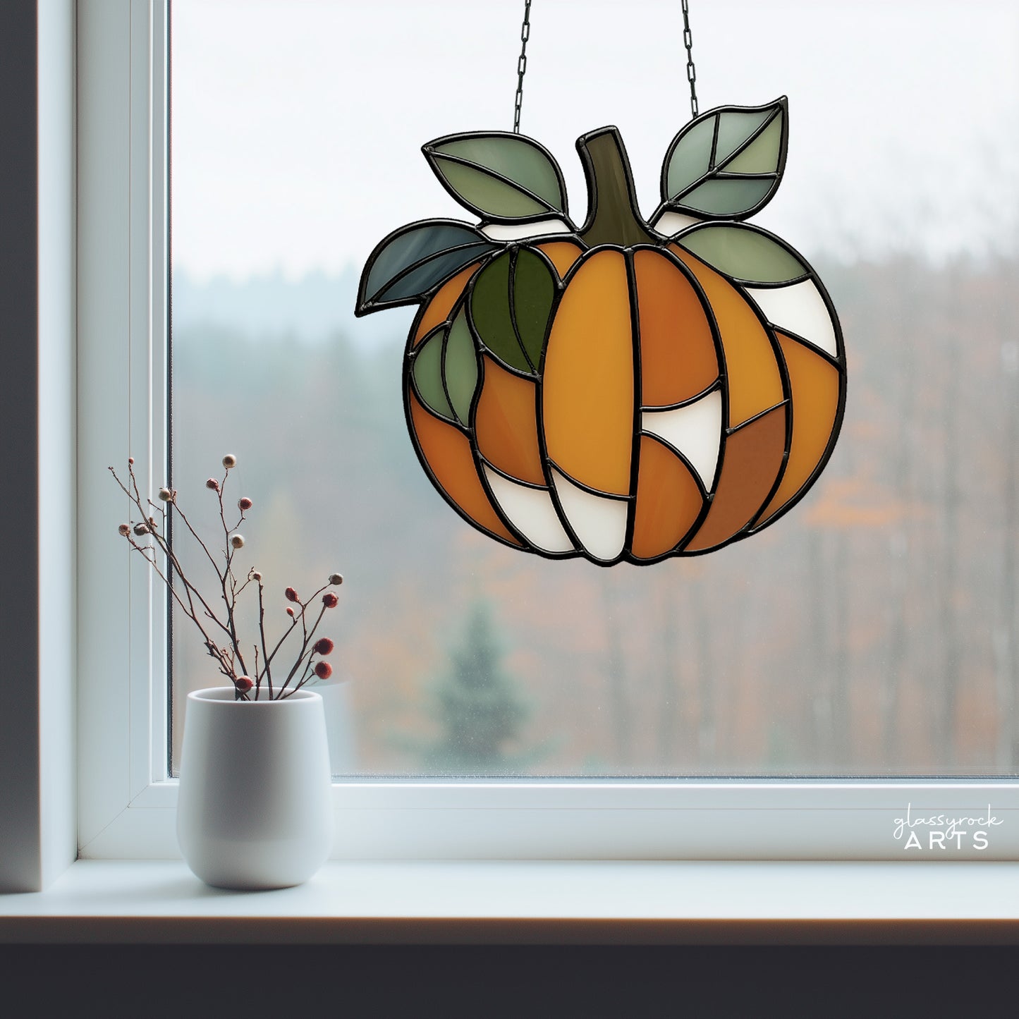 A leafy pumpkin suncatcher, hanging in a window with fall foliage outside. Inside there is a windowsill with some berries in a small white vase.