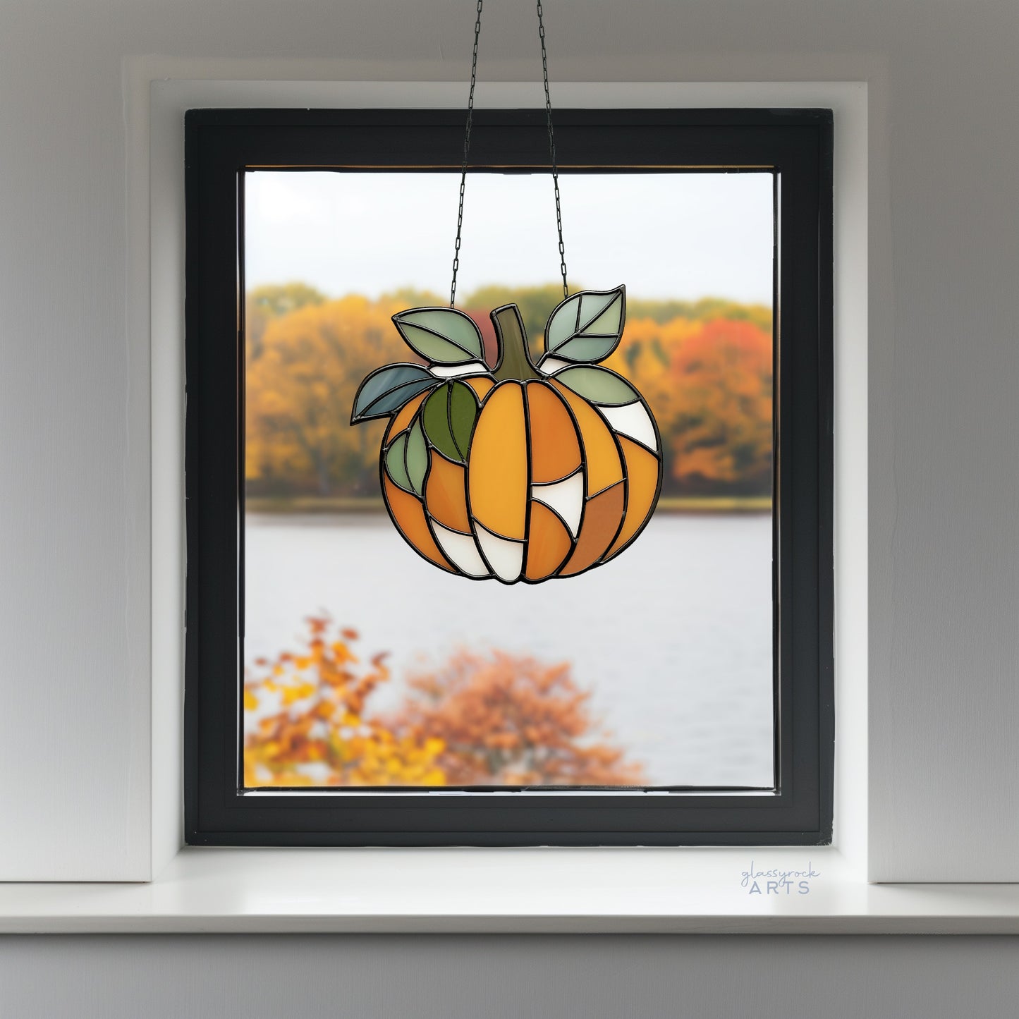 A leafy pumpkin suncatcher, hanging in a window with fall foliage outside. 