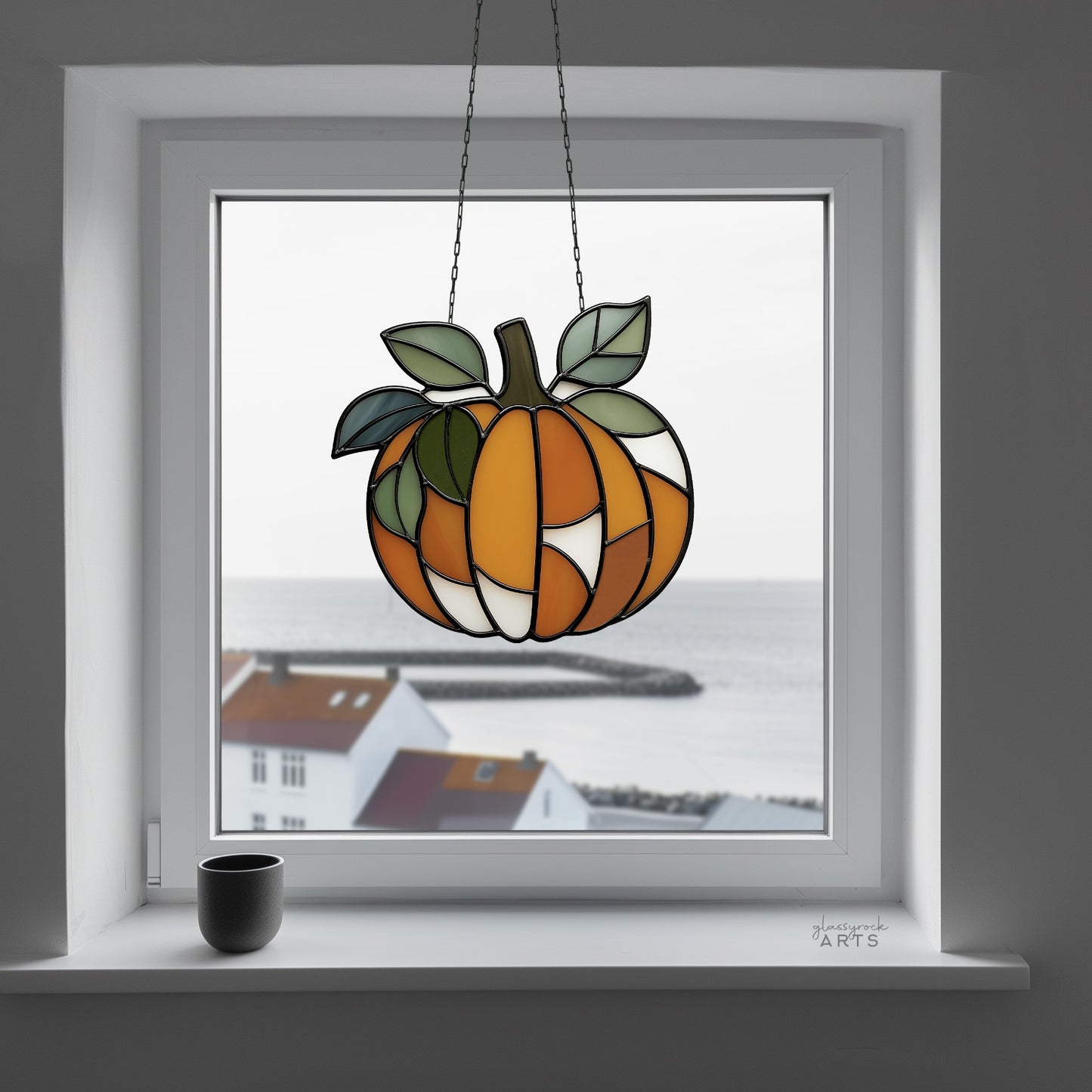 A leafy pumpkin suncatcher, hanging in a window with a harbor scene outside, and a small ceramic cup on the windowsill inside.