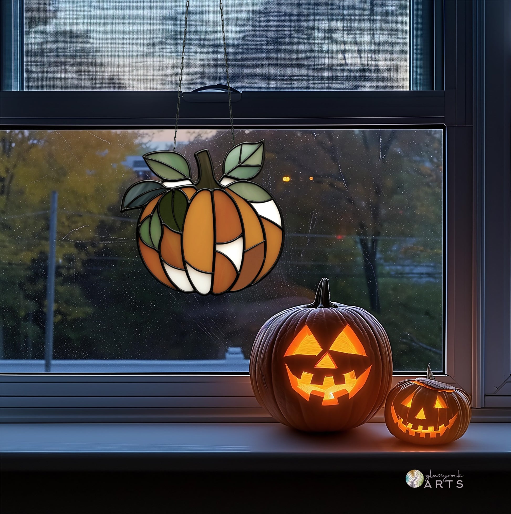 A leafy pumpkin suncatcher, hanging in a window with fall foliage outside at dusk. Inside there is a windowsill with two jack-o-lanterns.