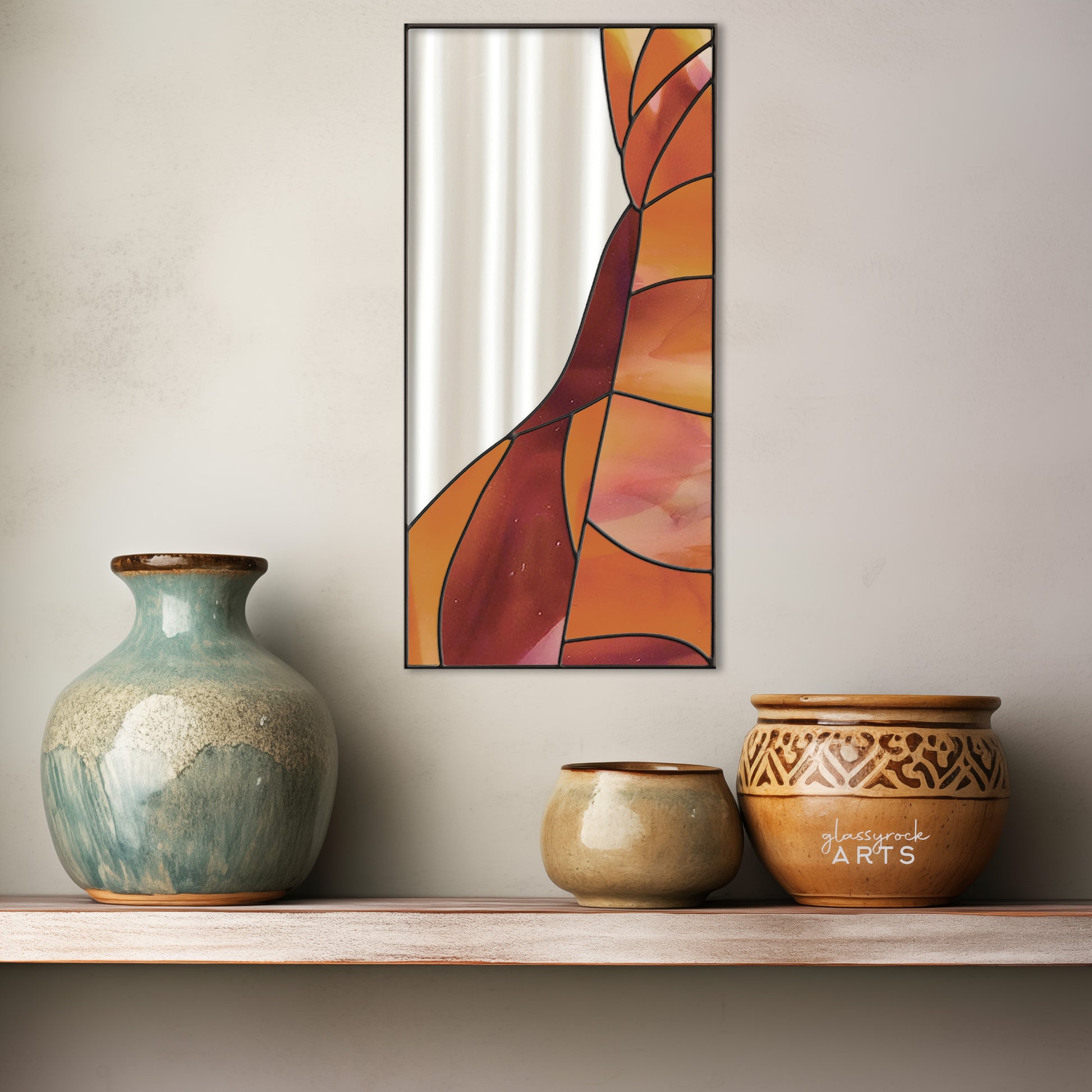 A rectangular mirror panel where the mirror glass occupies the upper left side while red and orange stained glass makes rock-like shapes along the rest of the piece. The panel is rectangular and tall. The panel is hanging on a wall above a shelf of beautiful handmade ceramic pottery.