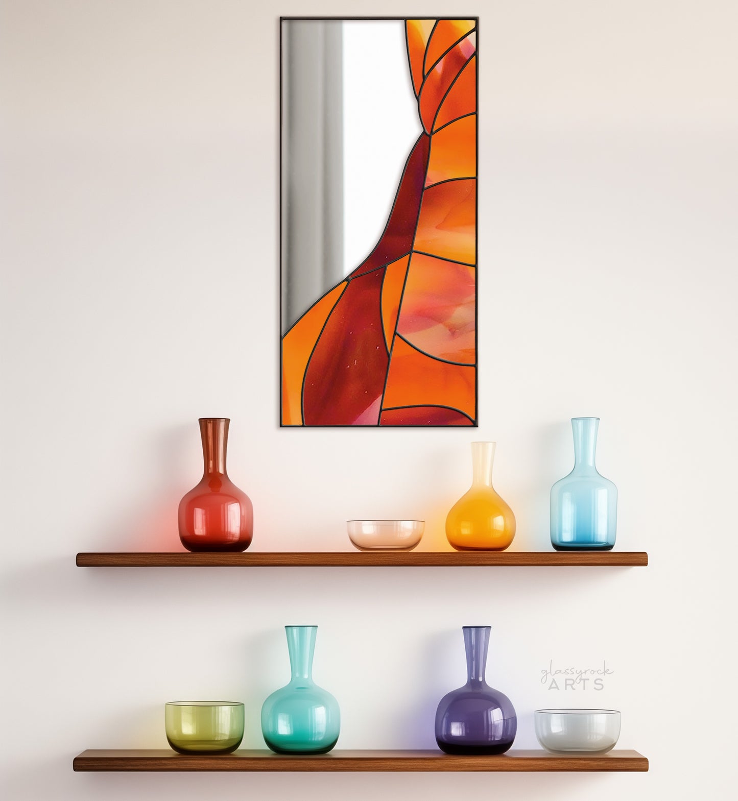 A rectangular mirror panel where the mirror glass occupies the upper left side while red and orange stained glass makes rock-like shapes along the rest of the piece. The panel is rectangular and tall. It hangs on a wall above two shelves of colorful glassware.