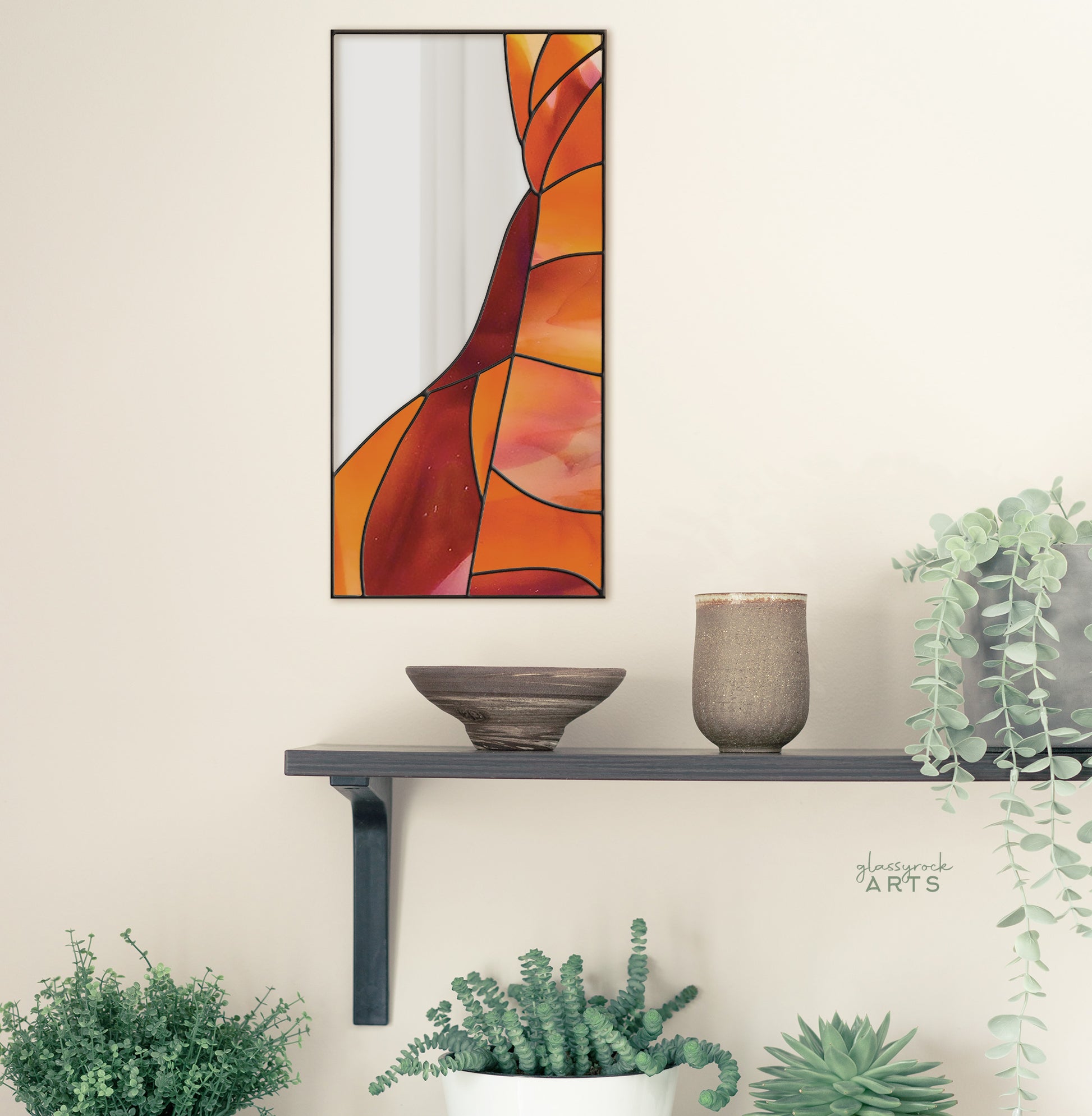 A rectangular mirror panel where the mirror glass occupies the upper left side while red and orange stained glass makes rock-like shapes along the rest of the piece. The panel is rectangular and tall. It hangs on a wall above shelves with plants and pottery.