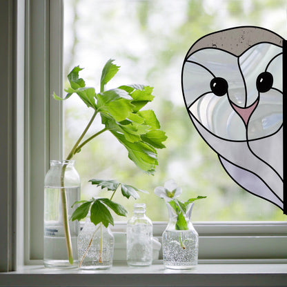 A vibrant Snowy Owl Stained Glass Pattern adorns the window, casting colorful hues. In front, three delicate glass bottles hold green leaves and a white flower, blending with the blurred greenery outside to merge art and nature.