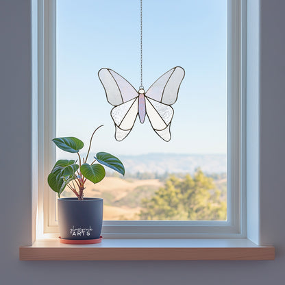 A simple stained glass butterfly made from 12 pieces of clear and iridescent glass, hanging in a window with a hilly background and a small plant on the windowsill.