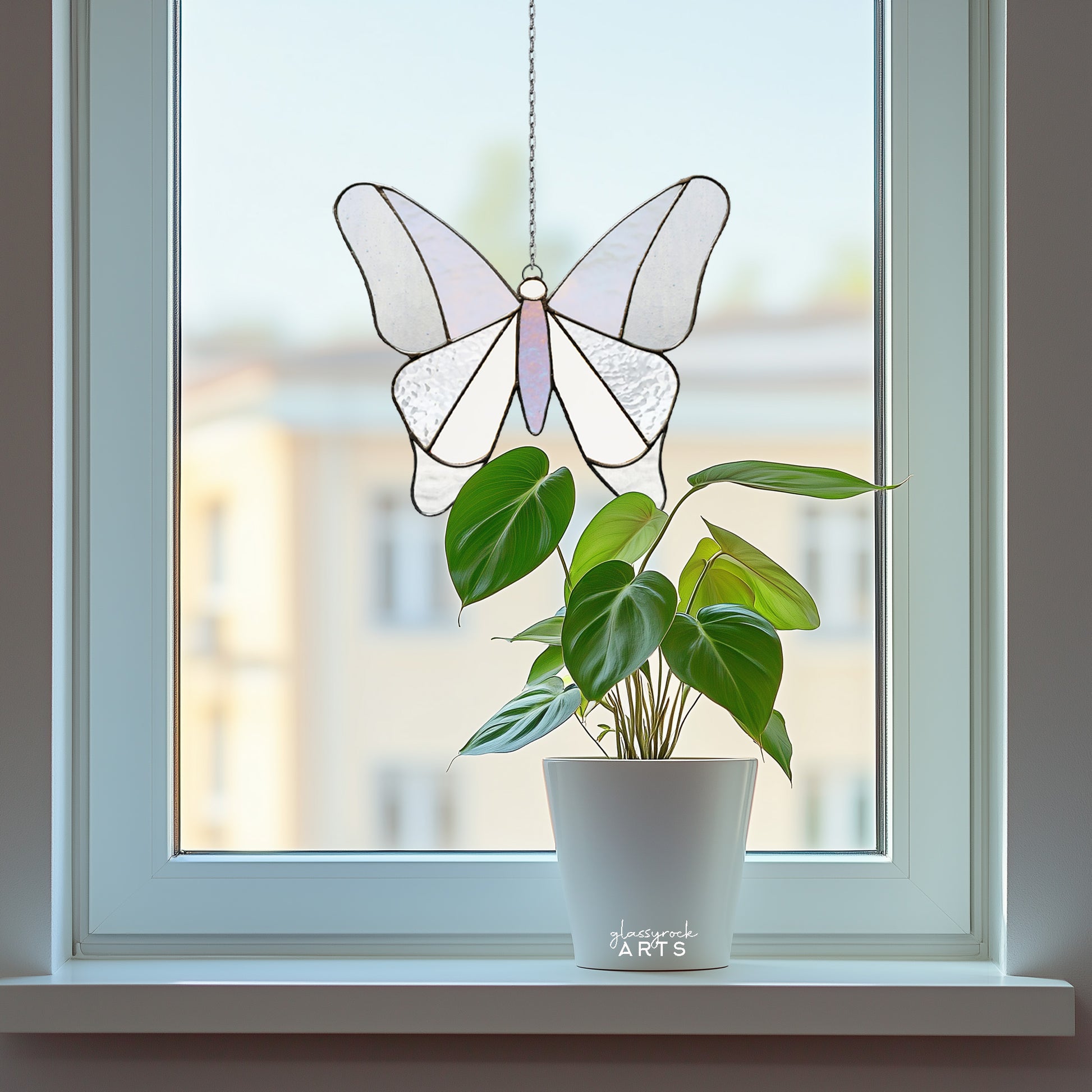 A simple stained glass butterfly made from 12 pieces of clear and iridescent glass, hanging in a window with an urban background and a plant on the windowsill.