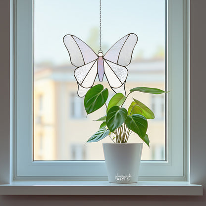 A simple stained glass butterfly made from 12 pieces of clear and iridescent glass, hanging in a window with an urban background and a plant on the windowsill.