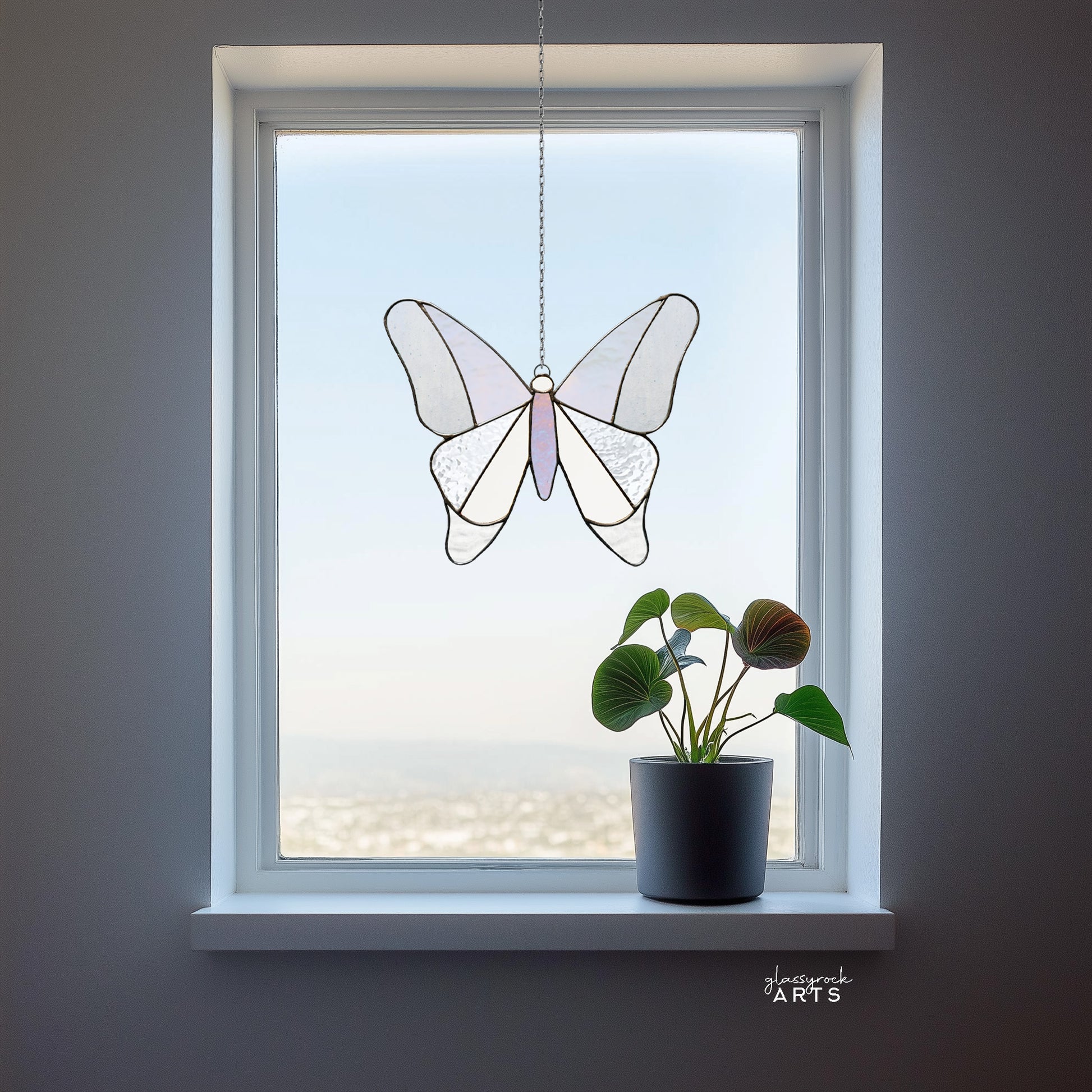 A simple stained glass butterfly made from 12 pieces of clear and iridescent glass, hanging in a window with a valley background and a small plant on the windowsill.