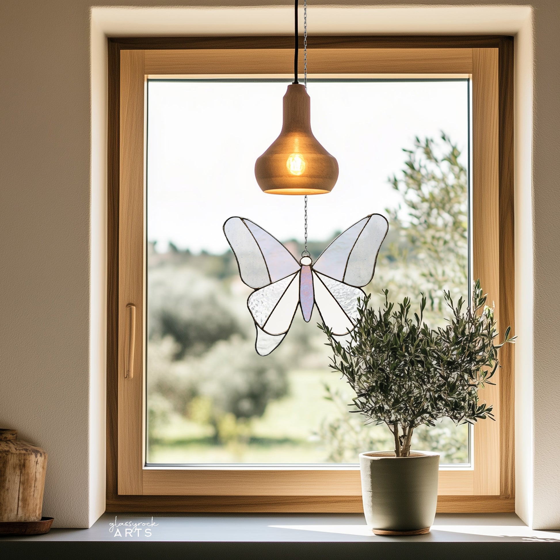 A simple stained glass butterfly made from 12 pieces of clear and iridescent glass, hanging in a window with a pastoral background and a potted small olive tree on the windowsill.