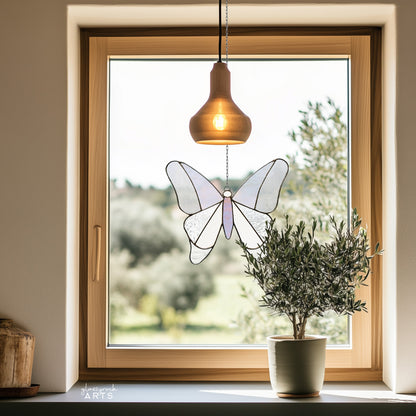 A simple stained glass butterfly made from 12 pieces of clear and iridescent glass, hanging in a window with a pastoral background and a potted small olive tree on the windowsill.