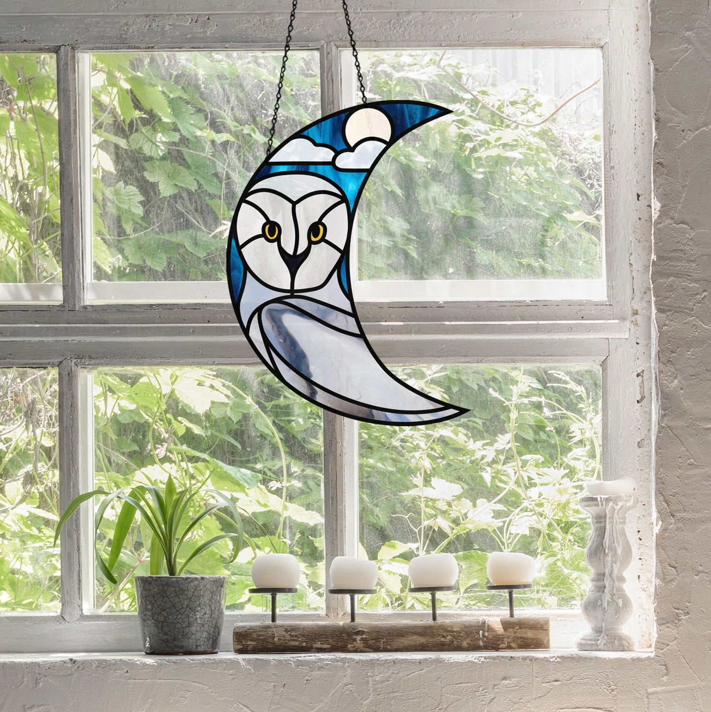 Stained glass owl design on a crescent moon hanging in front of a window with greenery outside.