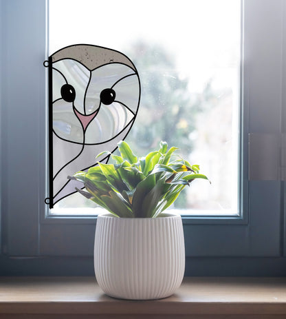 A white potted plant on a windowsill catches natural light beside a playful stained glass effect owl sticker. This owl, with its smiley face, is perfect for anyone considering the Snowy Owl Stained Glass Pattern digital download.