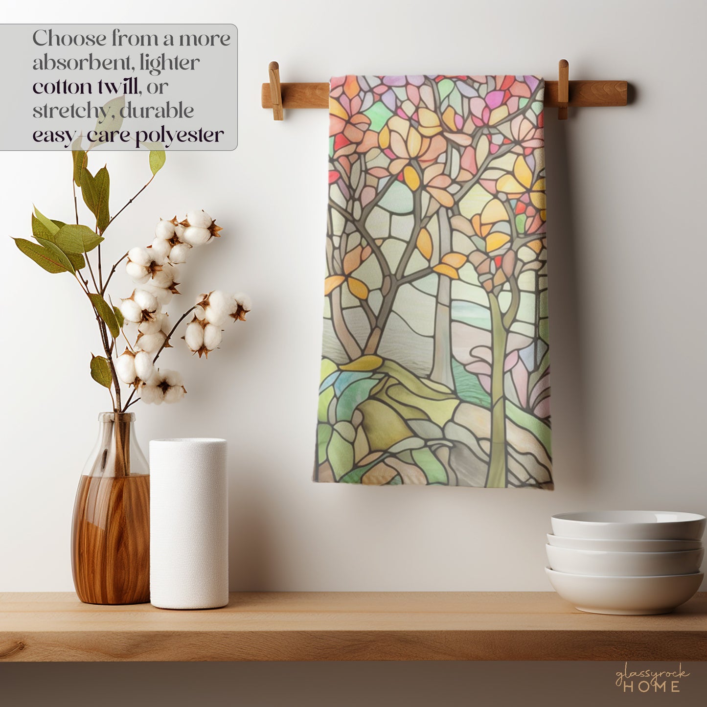 The Stained Glass Spring Forest Dish Towel with a vibrant tree pattern hangs on a wooden rack, paired with a cotton plant, white paper towels, and stacked bowls. Text reads: Opt for absorbent cotton twill or stretchy, durable polyester.