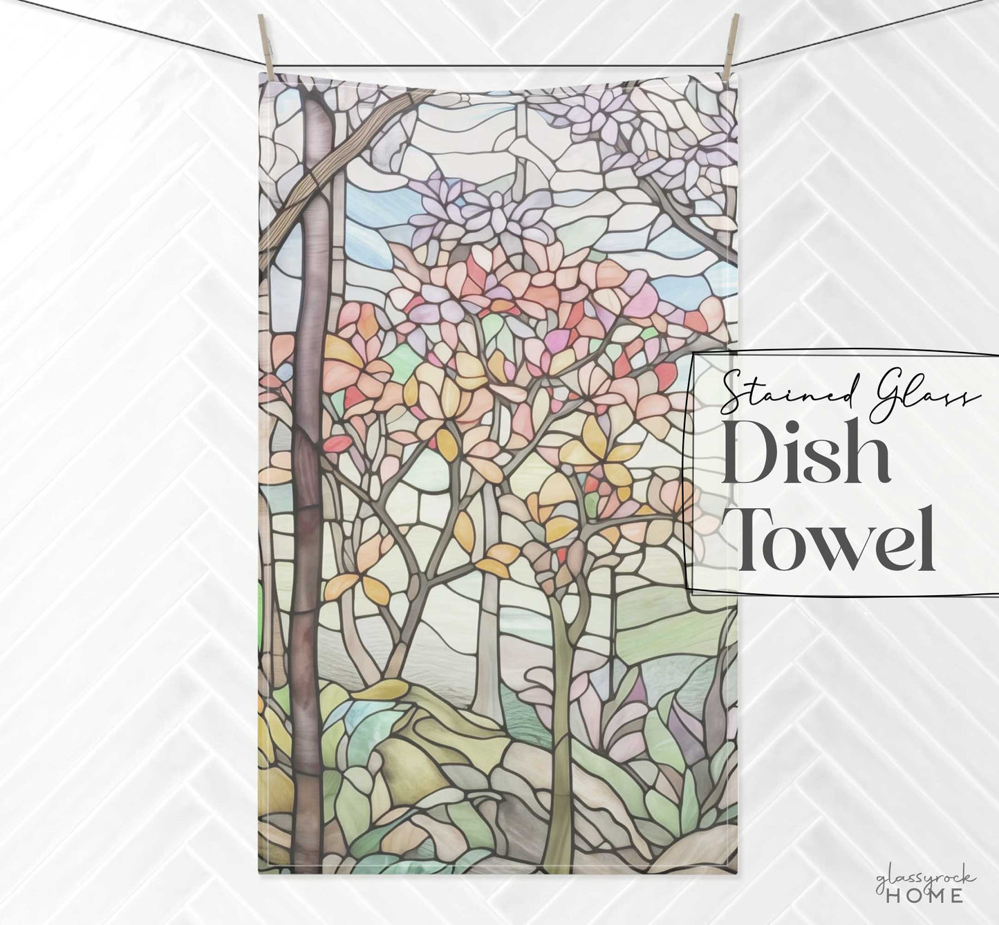 Against a textured white backdrop, the Stained Glass Spring Forest Dish Towel hangs, featuring a vibrant stained glass tree design with pink, orange, and purple foliage on a light blue sky background.