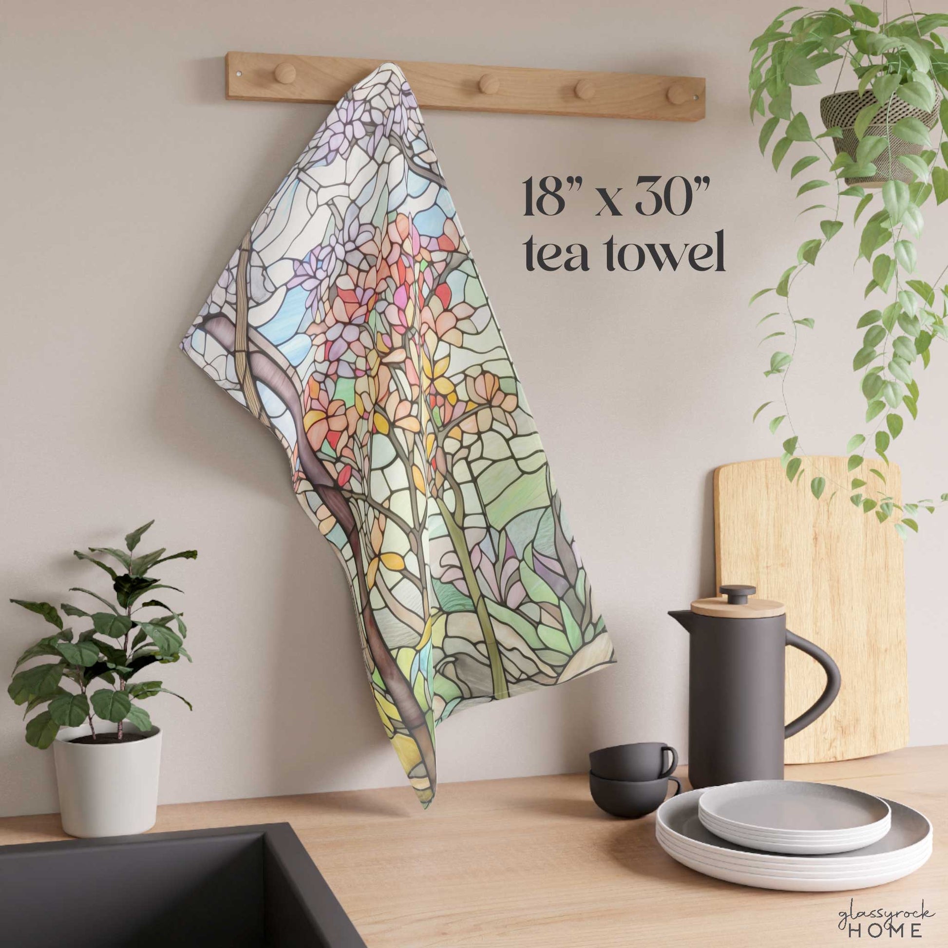 A colorful kitchen decor scene with an 18" x 30" Stained Glass Spring Forest Dish Towel featuring a floral design hanging on a wooden hook. Nearby are a potted plant, a coffee pot, a cup, and some plates.