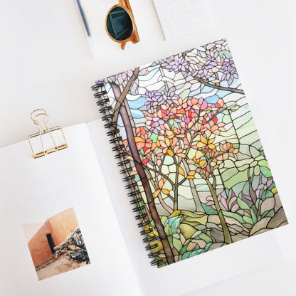 The Stained Glass Spring Forest Lined Spiral Notebook, with its vibrant tree and leaf design, is open on a white surface alongside a paper clip, sunglasses, and notepad. Ideal for journaling, it sits next to an open book with an orange building photo—a perfect stained glass gift!.