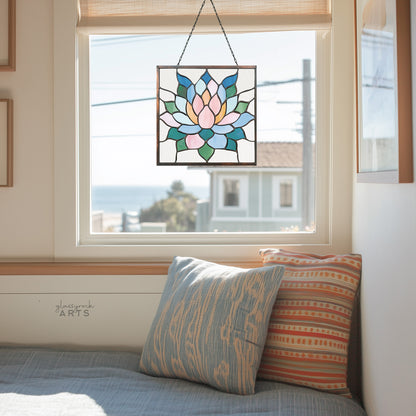 A beautiful square stained glass succulent panel, hanging in a window with a California coastal town background and a window seat with two pillows.