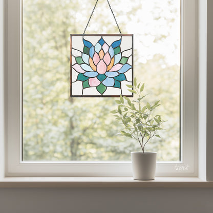 A beautiful square stained glass succulent panel, hanging in a window with a green trees background and a small plant on the windowsill.