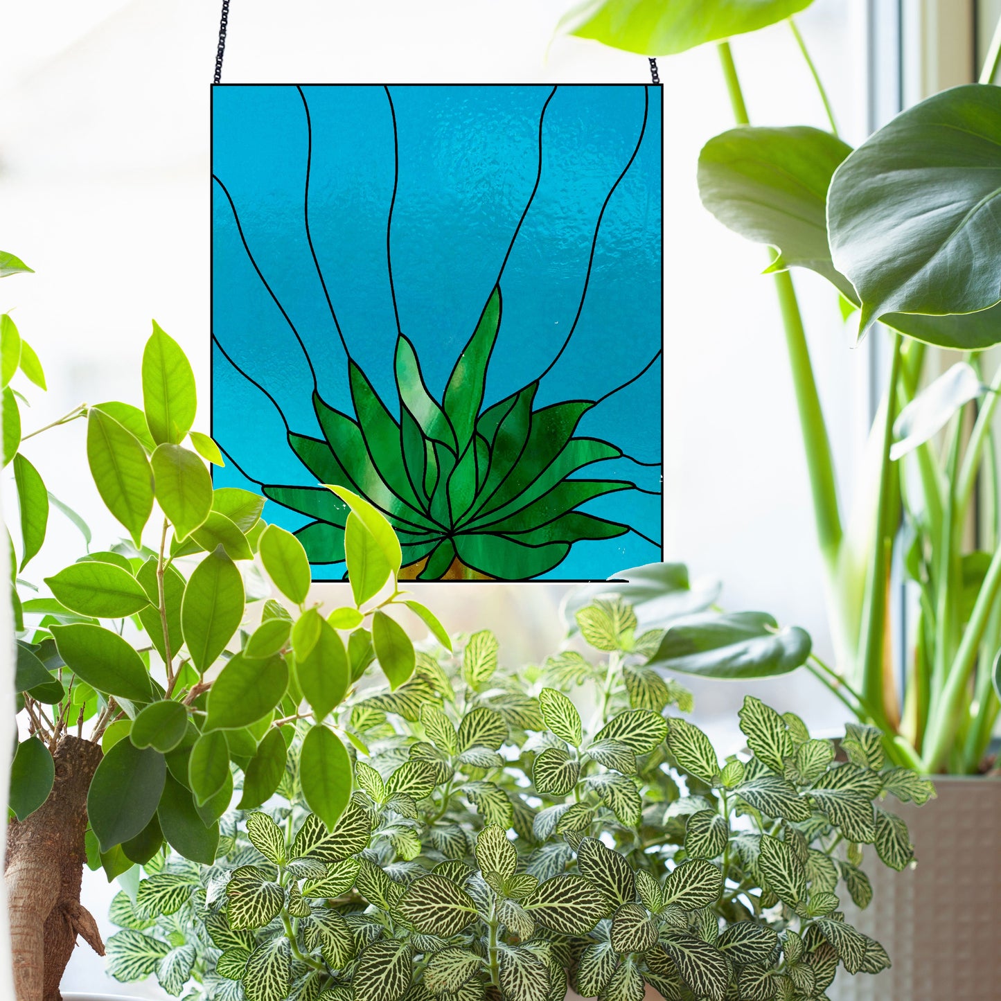 The Three Succulent Stained Glass Panel Patterns artwork features a green plant design, hanging in front of a window. Surrounded by real potted plants with varied leaf shapes and textures, it offers a harmonious blend of art and nature—a perfect unique decorative piece.