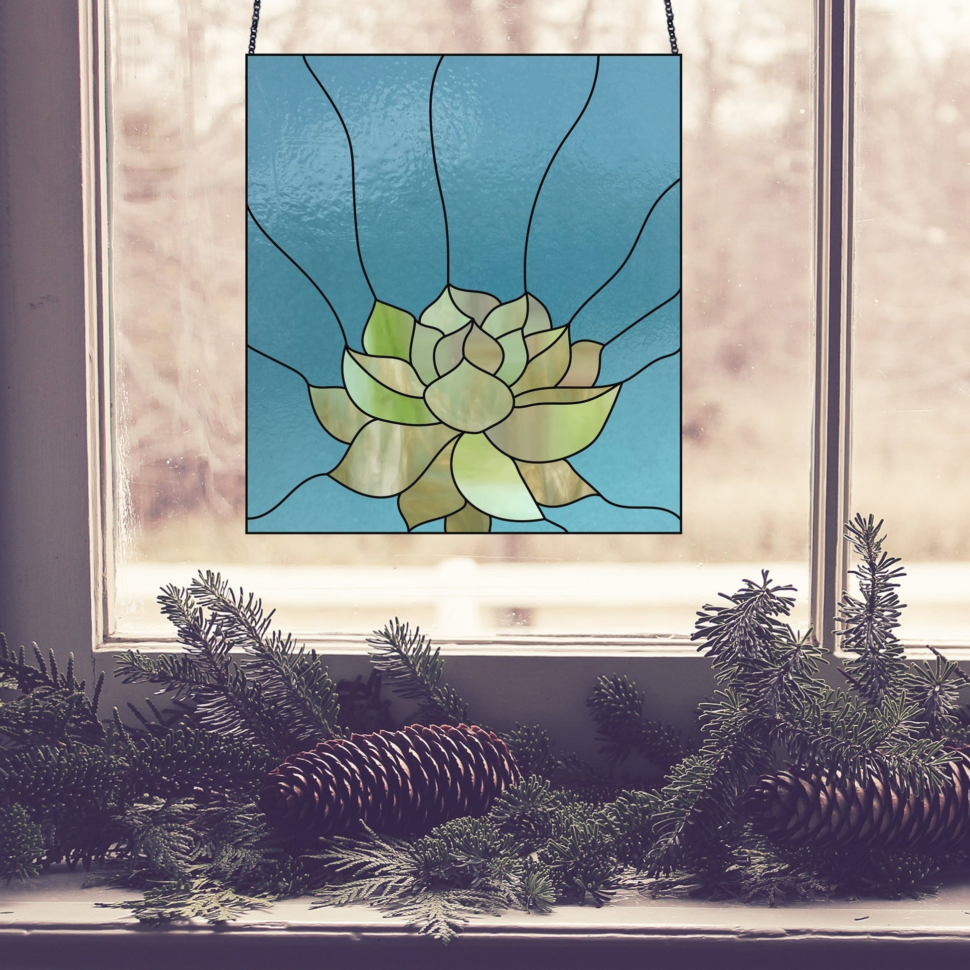 A Three Succulent Stained Glass Panel Patterns piece showcasing a green floral design hangs in a window, while pine branches and cones adorn the ledge below. The softly blurred background hints at trees beyond.