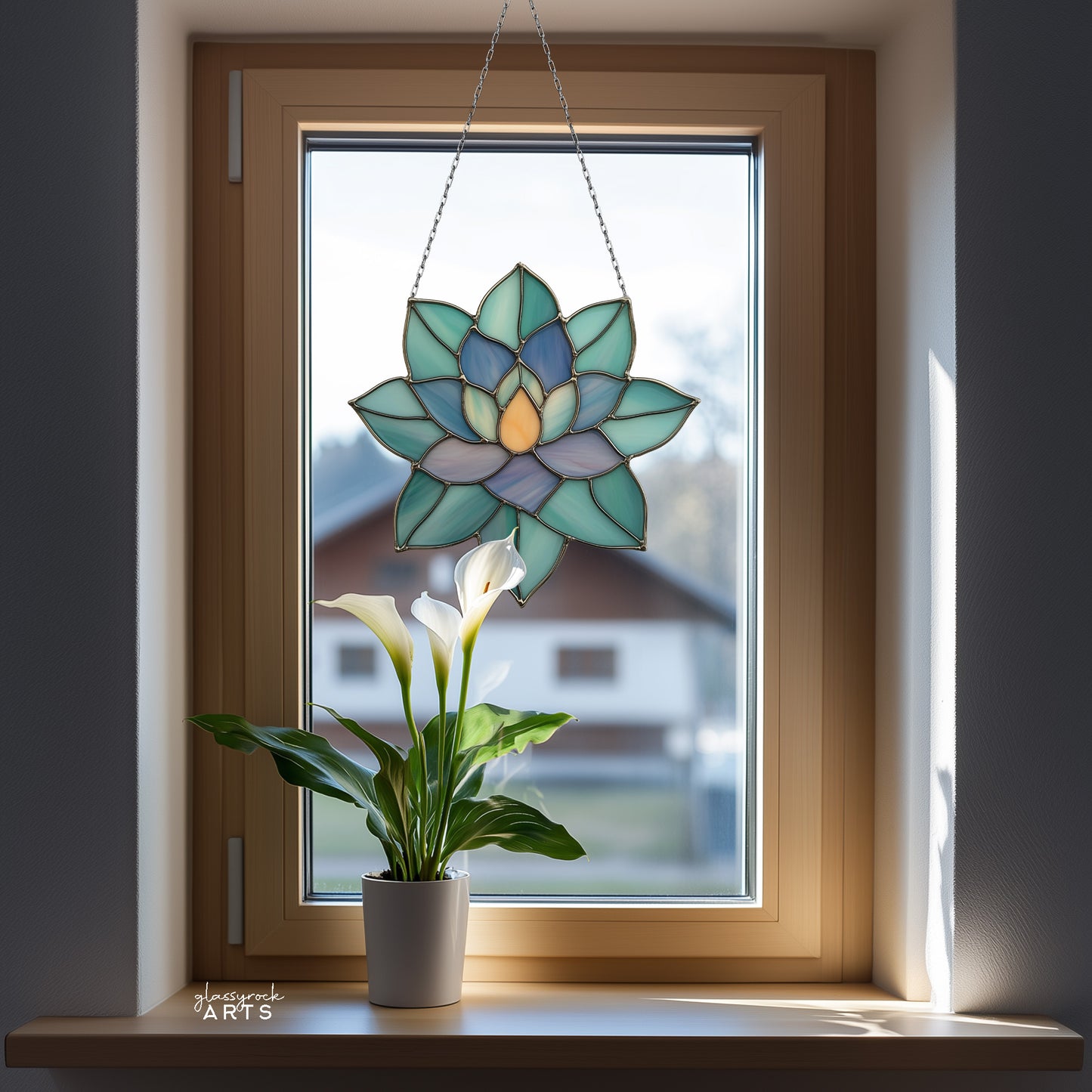 A colorful succulent suncatcher hanging in front of a window that has trees outside. There is a calla lily on the windowsill.