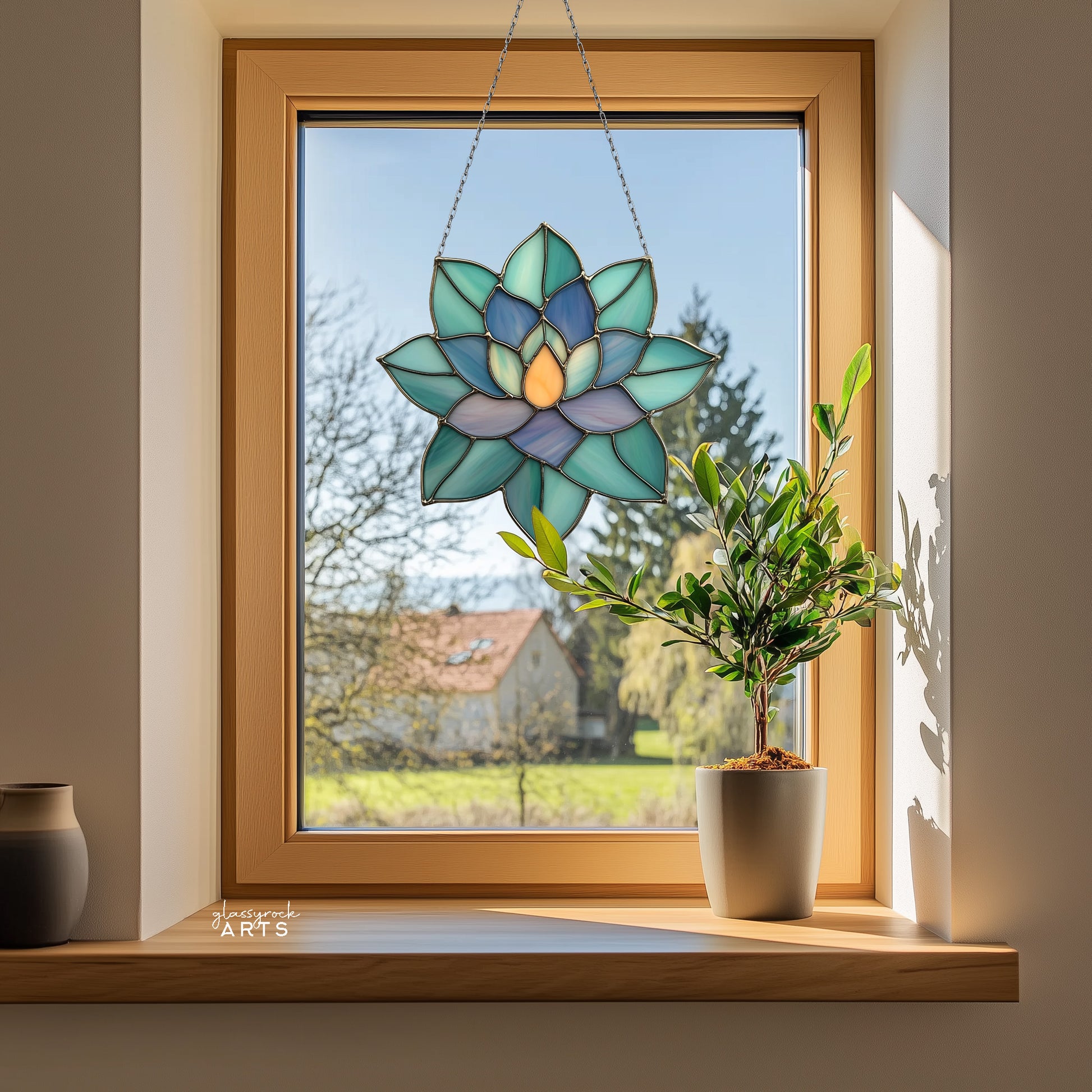 A colorful succulent suncatcher hanging in front of a window that has trees outside. There is a small plant on the windowsill.