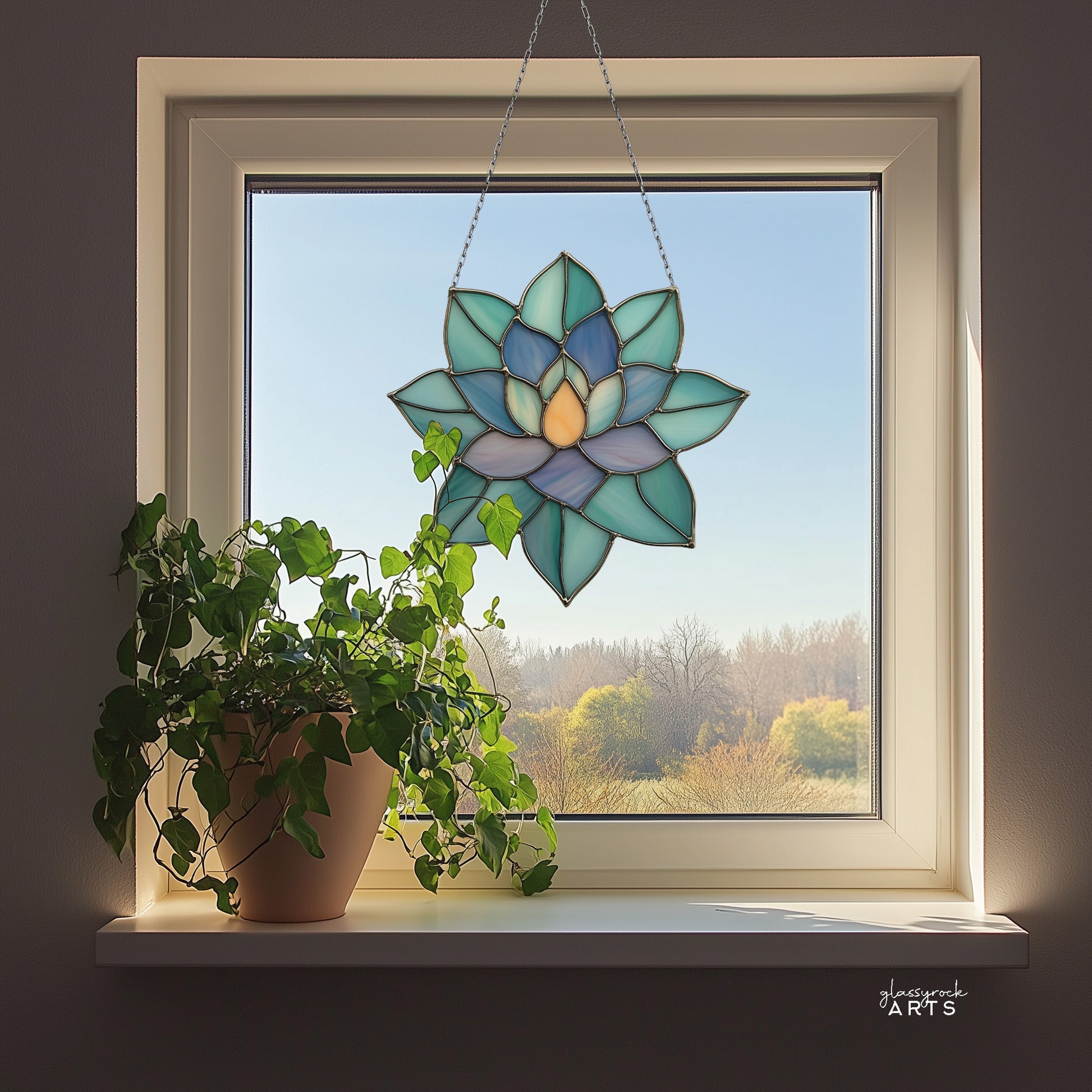 A colorful succulent suncatcher hanging in front of a window that has trees outside. There is a small plant on the windowsill.