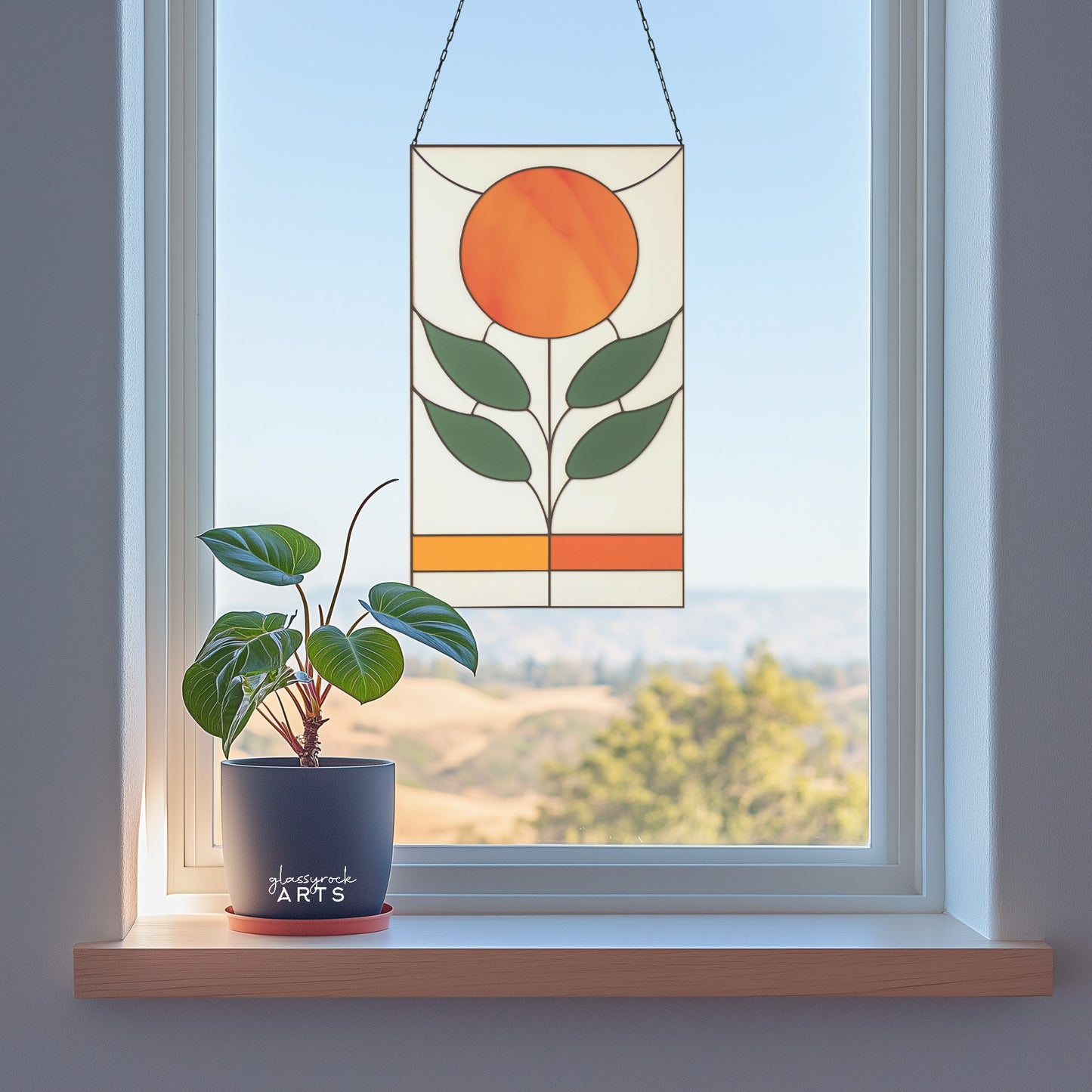 An orange, white, and green stained glass panel featuring a sun as the flower and four green leaves. The panel is rectangular and tall and hangs in a window with mountains behind.