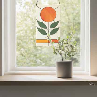 An orange, white, and green stained glass panel featuring a sun as the flower and four green leaves. The panel is rectangular and tall and hangs in a window with trees behind.