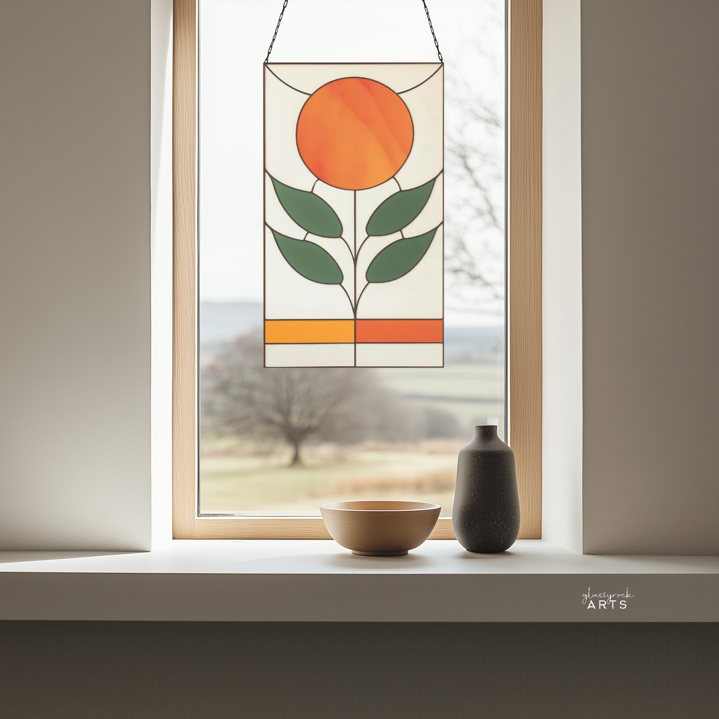 An orange, white, and green stained glass panel featuring a sun as the flower and four green leaves. The panel is rectangular and tall and hangs in a window with trees behind.