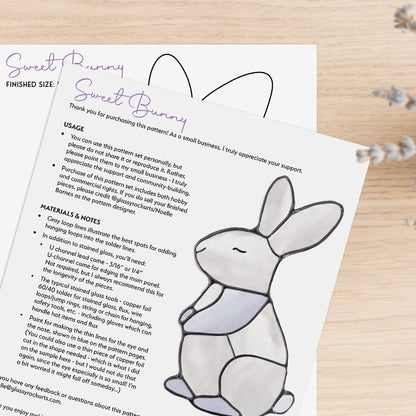 The Bunny Stained Glass Pattern is a digital download resting on a wooden surface, featuring a drawing of a seated bunny in gray and purple. Additional text includes usage instructions and material notes, capturing the charm of stained glass artistry.