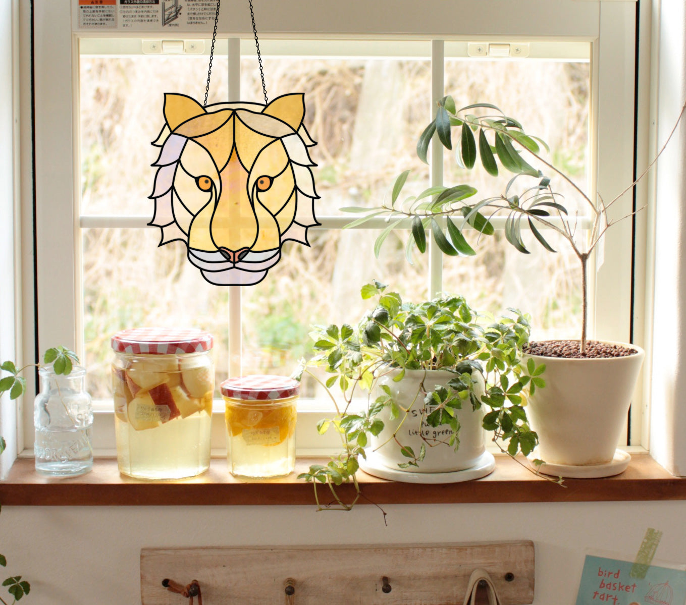 A sunny windowsill adorned with the Tiger Eyes Stained Glass Pattern features potted plants, jars with red-checked lids, a small bottle, and a note. Wall hooks are visible below the wooden ledge.