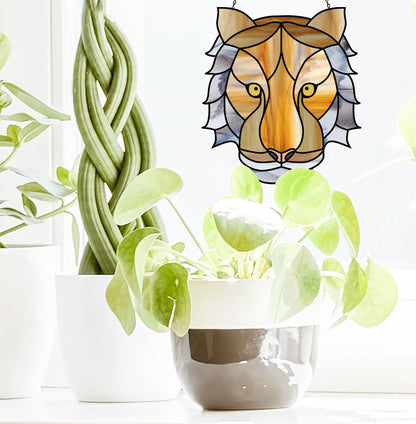 A sunlit room houses two potted plants: one with twisted stems, the other with broad, round leaves. Above them hangs the Tiger Eyes Stained Glass Pattern, casting colorful patterns and a lions face motif across the room.