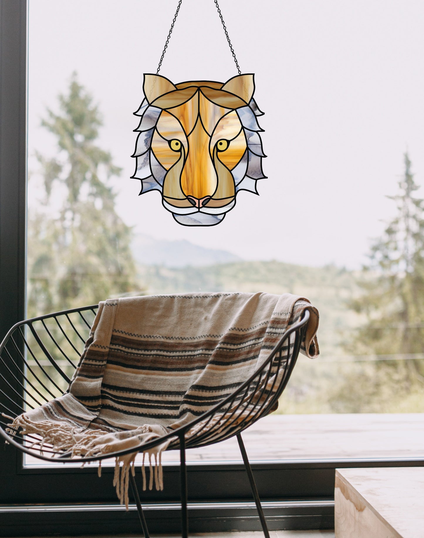 A black wire chair with a striped throw sits in front of a large window. The Tiger Eyes Stained Glass Pattern hangs there, adding charm. Trees and hills stretch out beyond the window.