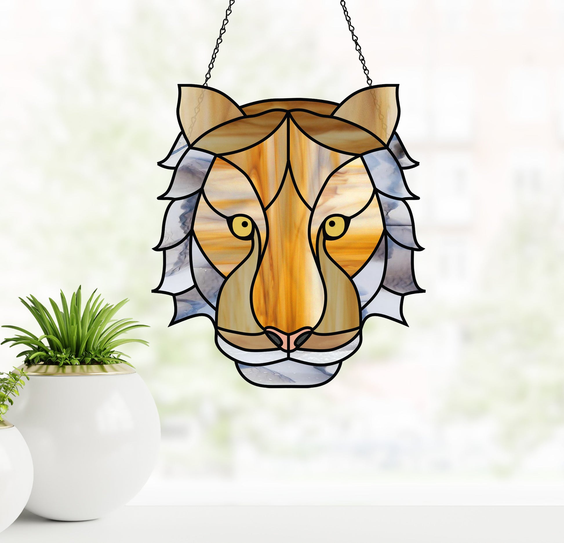 The Tiger Eyes Stained Glass Pattern, featuring a boho tiger face in yellow, brown, and gray hues, hangs from a chain against a blurred backdrop. Nearby are two white pots with green plants, capturing natures elegance.