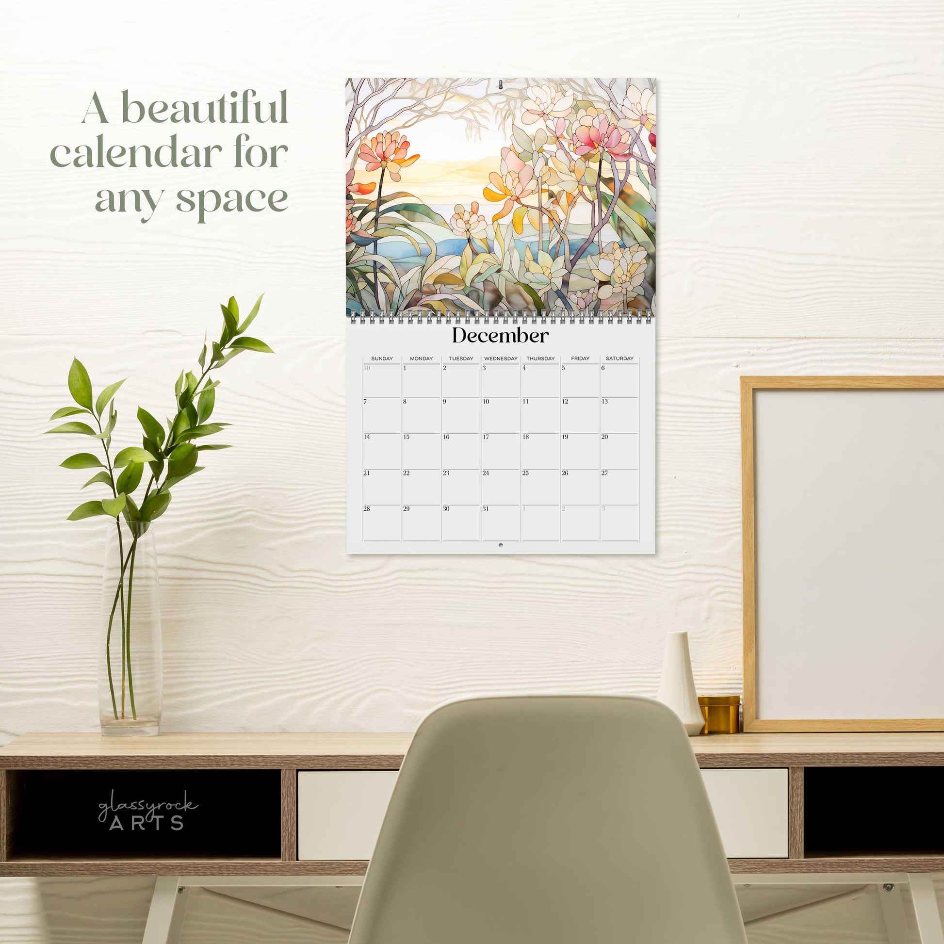 A wall-mounted 2025 Wall Calendar - Stained Glass Tropical Blooms featuring vibrant tropical flower illustrations above a desk with a plant, a framed picture, a chair, and a cup. The calendar’s high-quality paper complements the serene workspace perfectly.