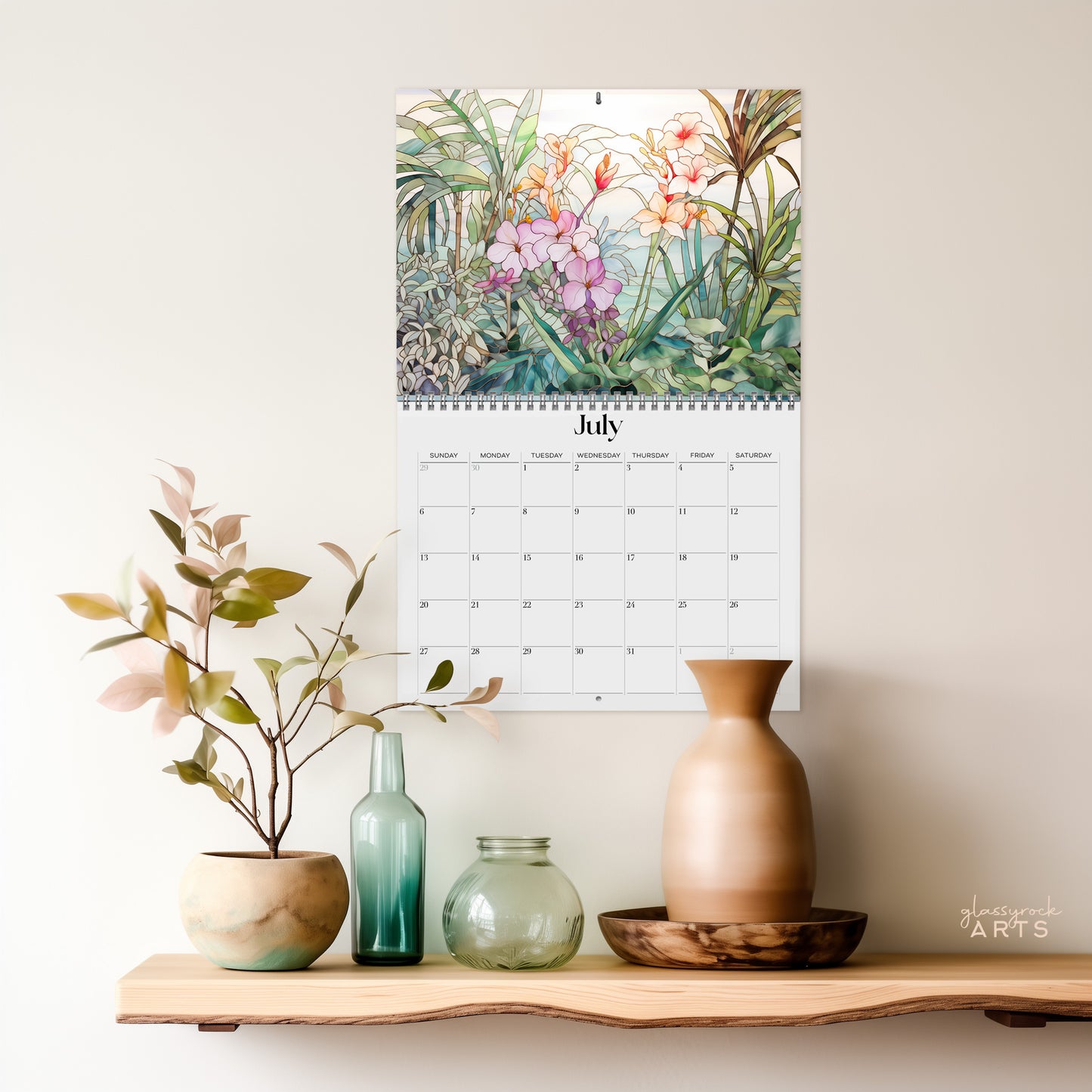 A "2025 Wall Calendar - Stained Glass Tropical Blooms" displaying the month of July hangs above a shelf adorned with various decorative vases and a plant. The high-quality paper calendar features an illustration of orchids and greenery.