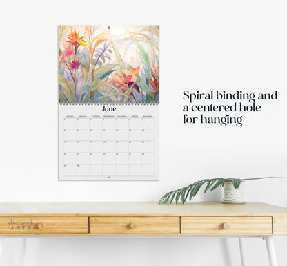 A 2025 Wall Calendar - Stained Glass Tropical Blooms, showcasing the month of June with intricate tropical flower illustrations at the top, spiral binding, and a centered hole for hanging. Below it is a wooden desk with a potted plant.