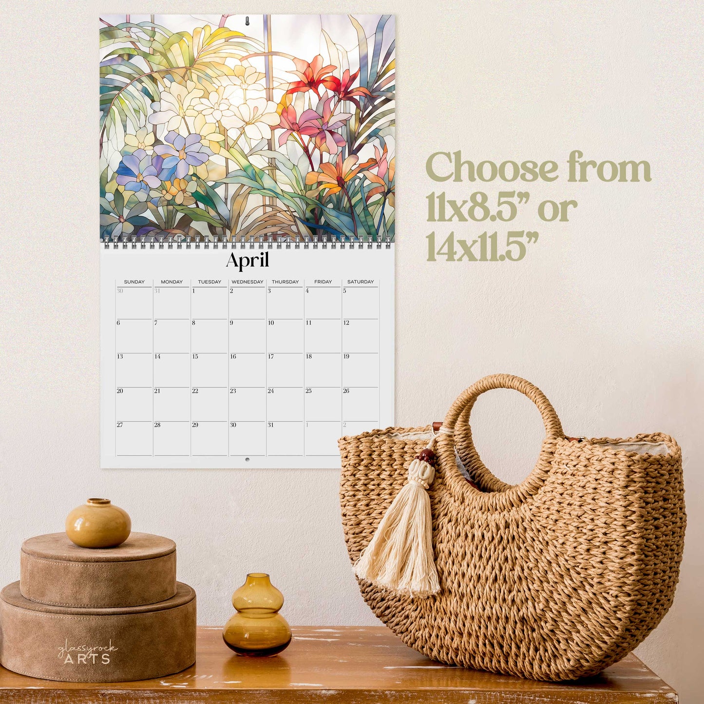 The 2025 Wall Calendar - Stained Glass Tropical Blooms for April hangs on a wall above a wooden surface adorned with a basket, a candle, and two stacked boxes. The high-quality paper showcases stunning tropical flower illustrations in stained glass designs. Text on the wall provides various size options for the calendar.