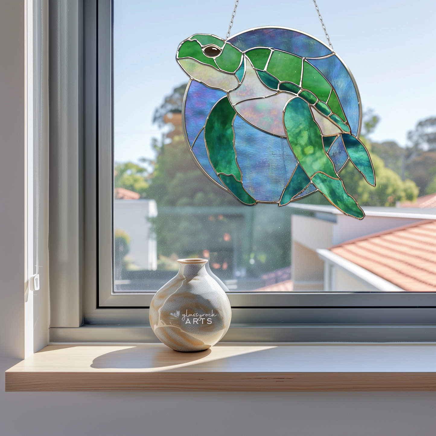 A Round Stained Glass Turtle Pattern sun catcher hangs in the window, casting colorful reflections. Below, a round ceramic vase with artistic patterns sits on the wooden windowsill, while rooftops and blurred trees are visible in the background.