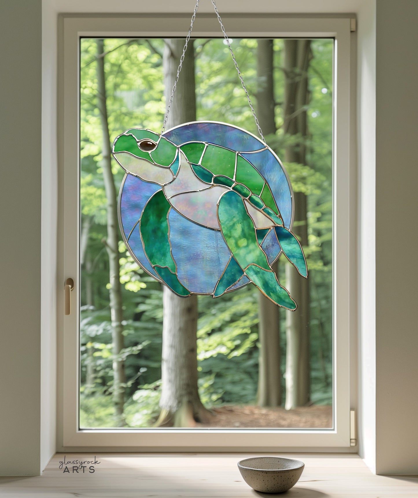 The Round Stained Glass Turtle Pattern hangs gracefully from a chain, framing the lush forest view. Below, on the windowsill, a small bowl filled with decorative gravel enhances the serene turtle decor.